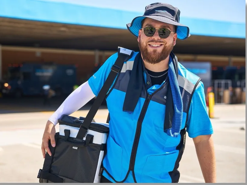 A delivery associate in an Amazon-branded safety vest carrying a delivery bag outside a delivery station